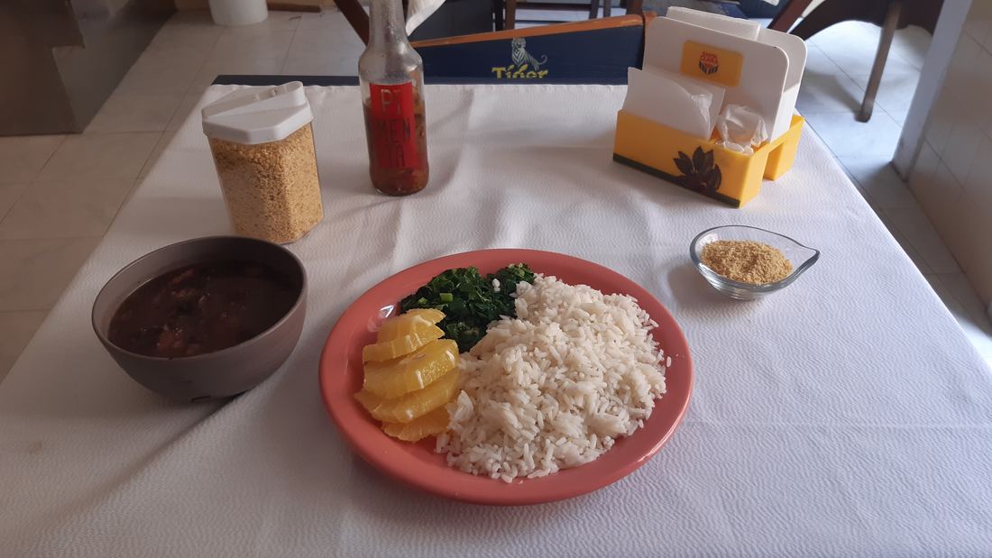 A plate of feijoada with rice, greens and orange slices on a restaurant table