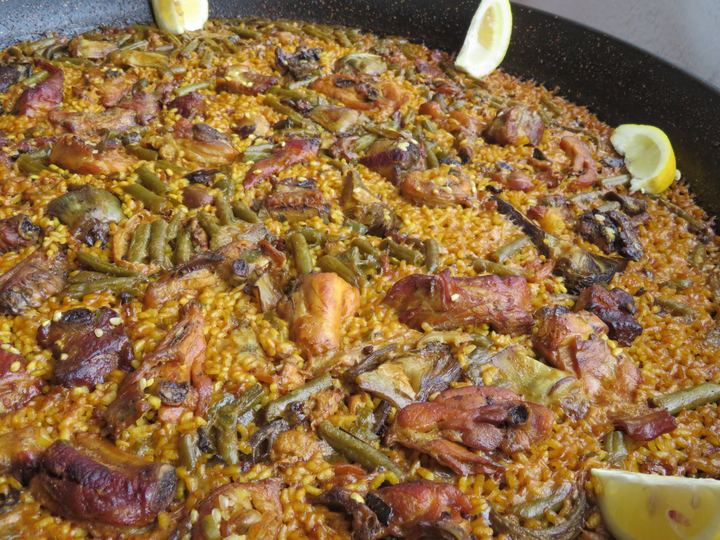 A wide pan of paella valenciana with meat, green beans and lemon wedges