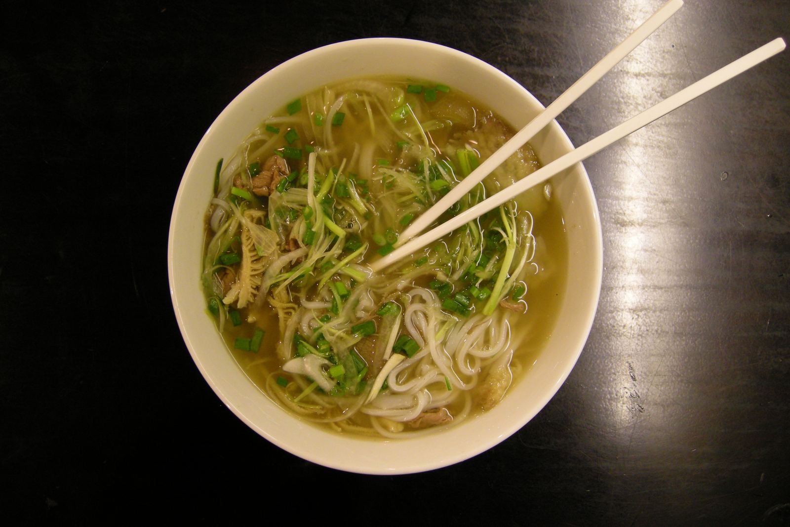 A bowl of pho with rice noodles, herbs and chopsticks on a dark table